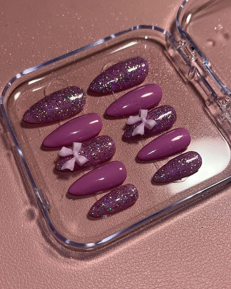 Purple almond press-ons with glitter and small white bows in a clear case